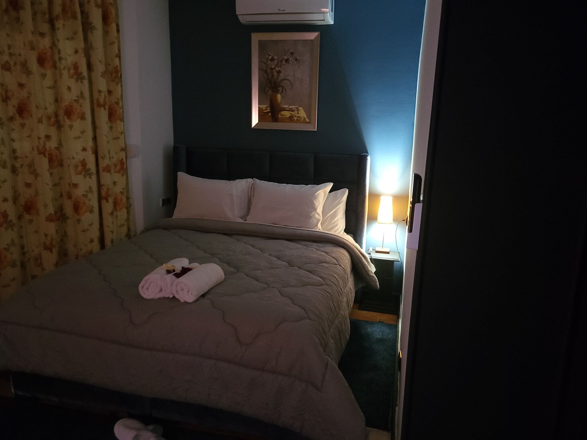 Una cama bien hecha con colcha color beige, dos almohadas y toallas enrolladas. Encima de la cama, hay un cuadro en la pared.