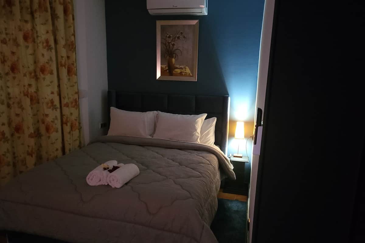 A neatly made bed with a beige quilt, two pillows, and rolled towels on the bed. There is a framed picture on the wall above the bed.