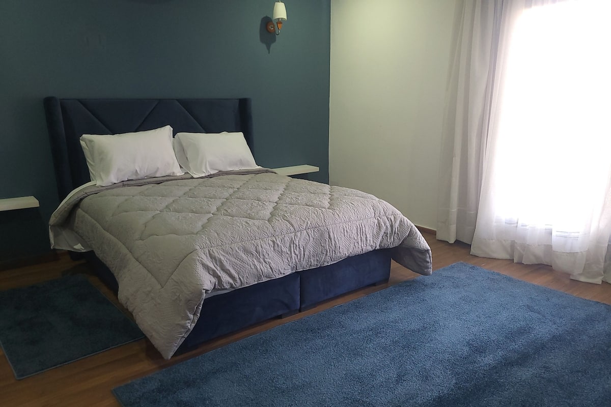 A bedroom with a bed, a nightstand, a wall-mounted lamp, and a window with curtains.