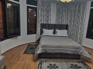 Une chambre à coucher avec un grand lit, une tête de lit et deux fenêtres équipées de stores.