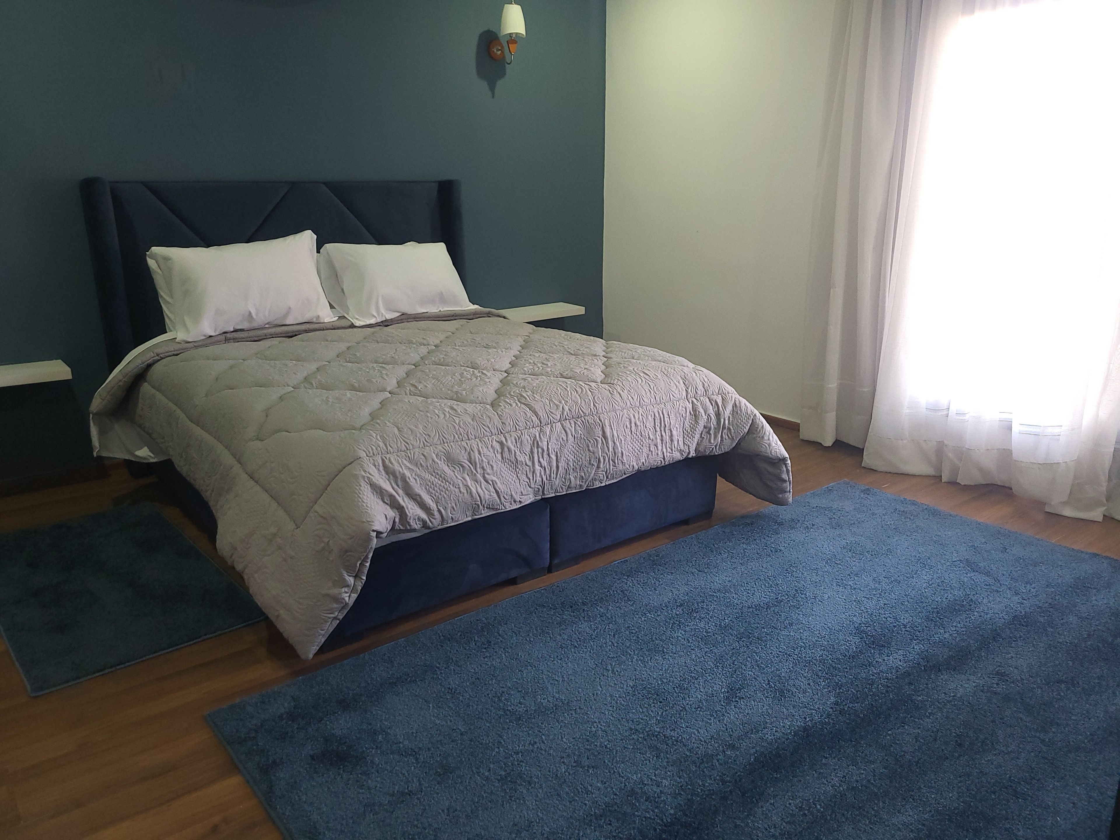 A bedroom with a bed, a nightstand, a wall-mounted lamp, and a window with curtains.