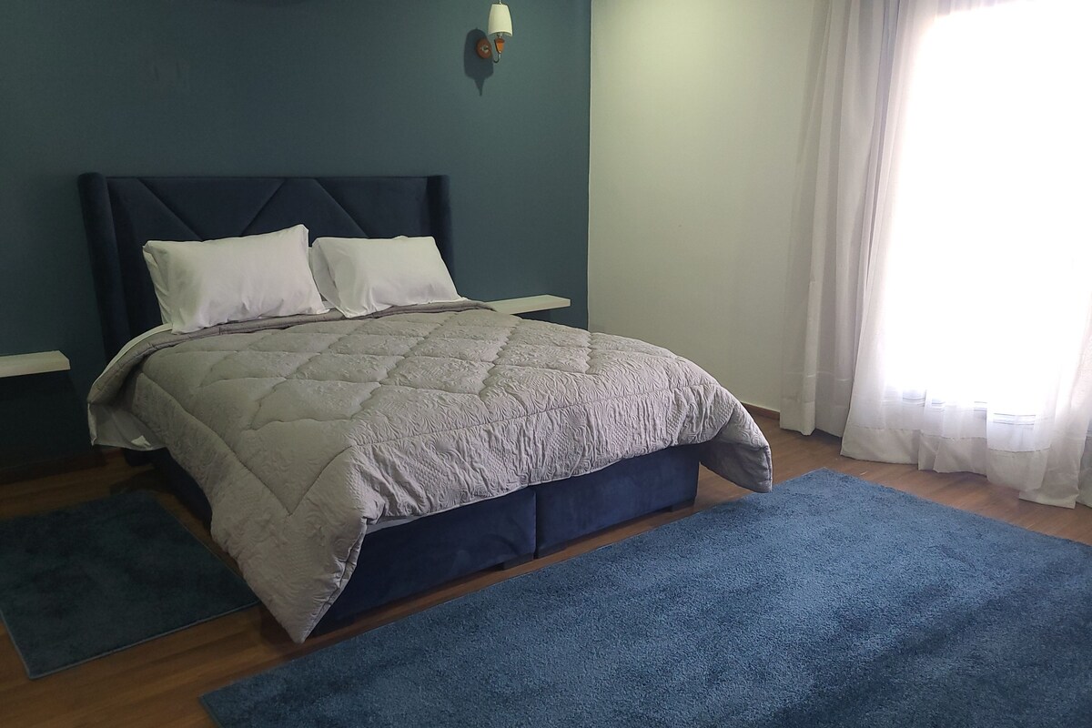 Une chambre à coucher avec un lit, une table de chevet, une lampe fixée au mur et une fenêtre avec des rideaux.