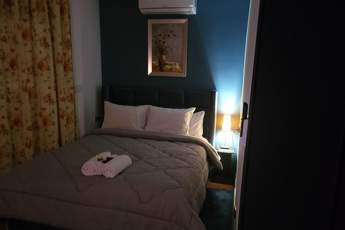 Un lit bien fait, recouvert d’une courtepointe beige, avec deux oreillers et des serviettes enroulées posées sur le lit. Un tableau encadré est accroché au mur, au-dessus du lit.