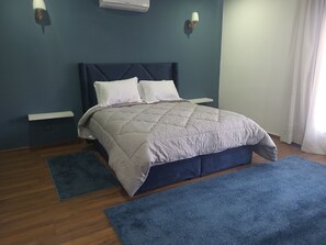 Une chambre à coucher comprenant un lit, deux tables de chevet, un climatiseur fixé au mur et une fenêtre avec des rideaux.
