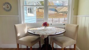 A dining area with a round table and two chairs, a window with a view of the outside, and a vase with flowers on the table. - Gorgeous cottage in the heart of the picturesque Huon Valley. (Castle Forbes Bay)