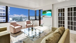 A modern living room with a glass coffee table, two armchairs, a sofa, and a large TV screen displaying a beach scene. - Beach Resort condo /921 (Miami Beach)