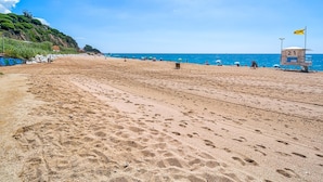 Une vaste plage de sable fin, parsemée d’empreintes, avec une cabine de sauveteur et un ciel bleu azur.