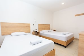 Una habitación de hotel con dos camas individuales, cabeceros de madera, ropa de cama blanca, una mesita de noche pequeña con una lámpara y una mesita auxiliar de madera.