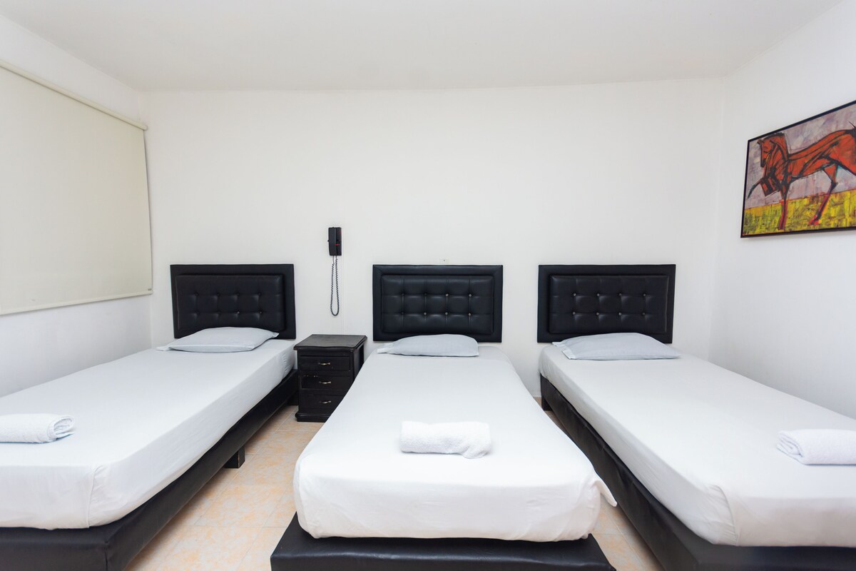 A hotel room with three single beds, a bedside table, a phone, and a painting on the wall.