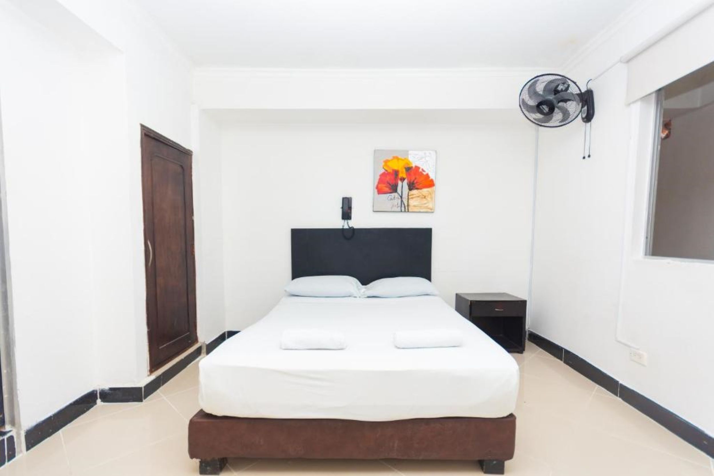 Habitación de hotel con una cama, una mesita de noche, un ventilador de pared y un cuadro.
