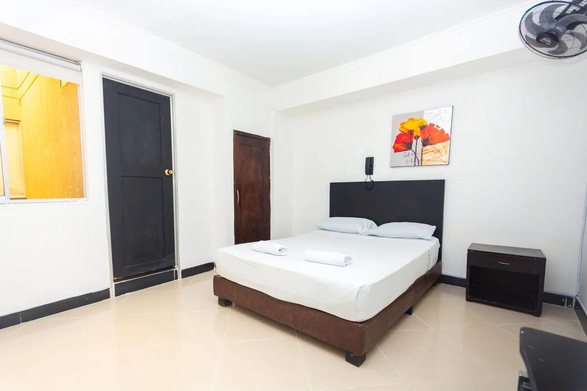 A hotel room with a bed, a bedside table, a door, and a wall-mounted fan.