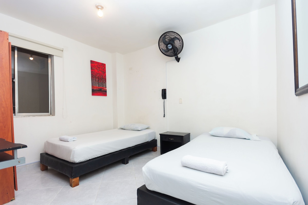 Una habitación con dos camas individuales, una mesita, una silla, un ventanal, un ventilador de pared y un cuadro abstracto rojo.