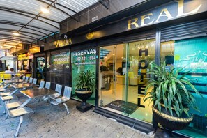 Een hotelgevel met terras, een receptiebalie en een grote potplant.
