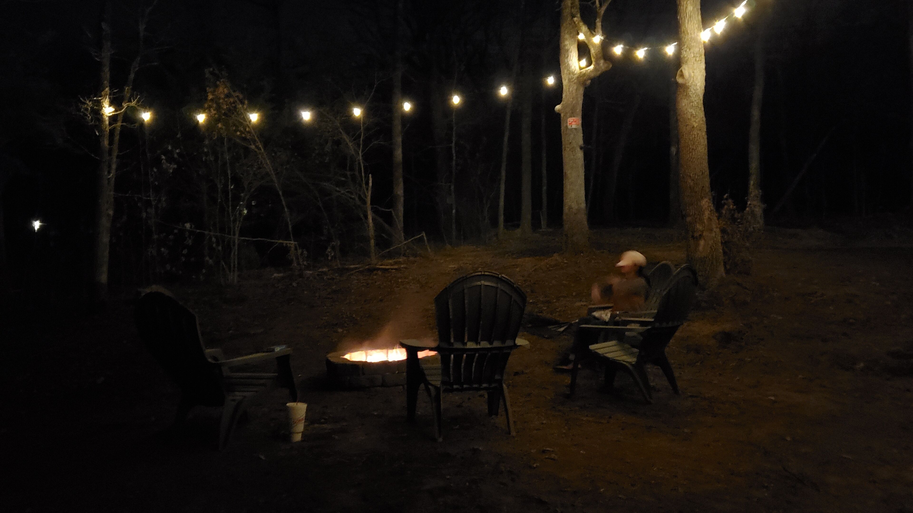 En campingplats med en eldstad, två stolar och ljusslingor i en skog under natten.