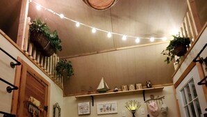 Una habitación acogedora con techo de madera, luces de adorno, un pequeño barco de vela decorativo, y varias plantas y adornos colgados en las paredes.