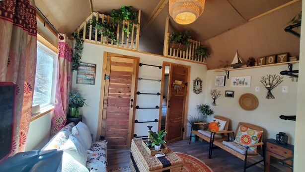 Una habitación acogedora con una escalera de madera, un sofá, una pequeña mesa con una planta y un sillón con un cojín colorido.