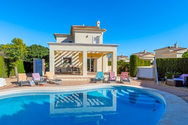 A poolside area with a clear blue pool, lounge chairs, and a building with a tiled roof.