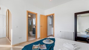 A bedroom with a bed, a bedside table, a mirror, and a view of a bathroom through an open door. - Villa Gran Sol 13 - PlusHolidays (Calpe)