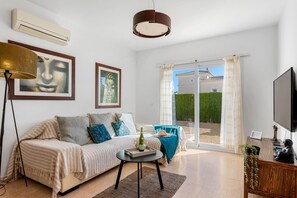 A modern living room with a sofa, a coffee table, and a television. There is a large window with curtains and a view of a building outside. - Villa Gran Sol 13 - PlusHolidays (Calpe)