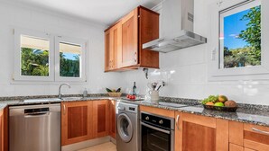 A kitchen with wooden cabinets, stainless steel appliances, a washing machine, and a window with a view of greenery. - Villa Gran Sol 13 - PlusHolidays (Calpe)