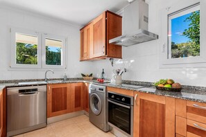 A kitchen with wooden cabinets, stainless steel appliances, a washing machine, and a window with a view of greenery.
