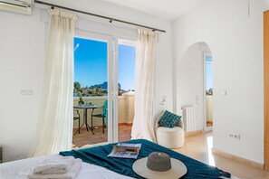 A bedroom with a bed, a window with curtains, a view of a balcony, and a chair with a pillow.