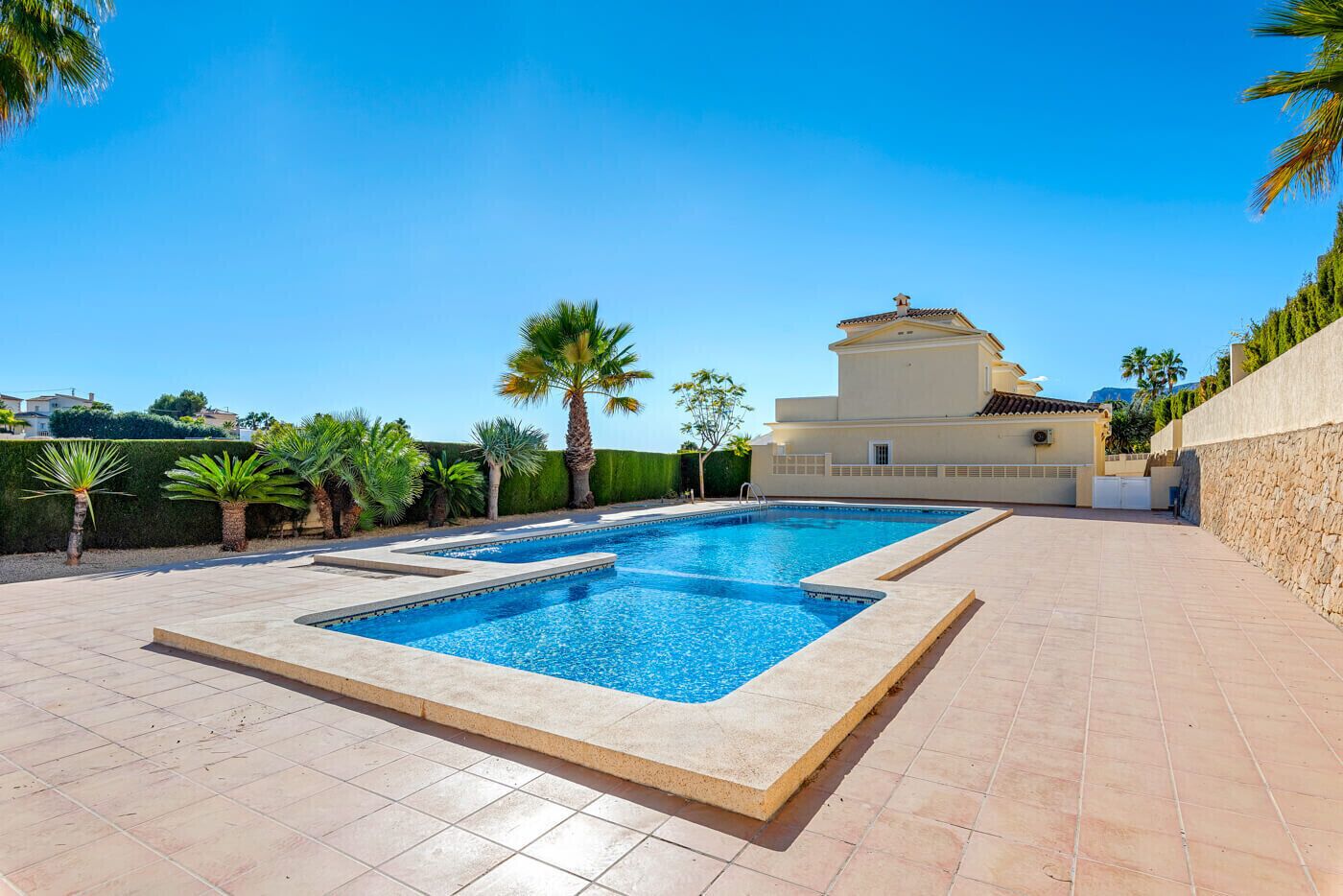 A rectangular swimming pool with a tiled deck, surrounded by palm trees and a building with a tiled roof.