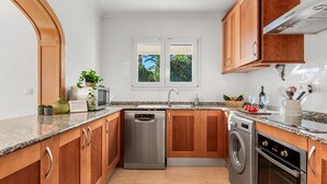 A modern kitchen with wooden cabinets, stainless steel appliances, and a window with a view of greenery. - Villa Gran Sol 13 - PlusHolidays (Calpe)