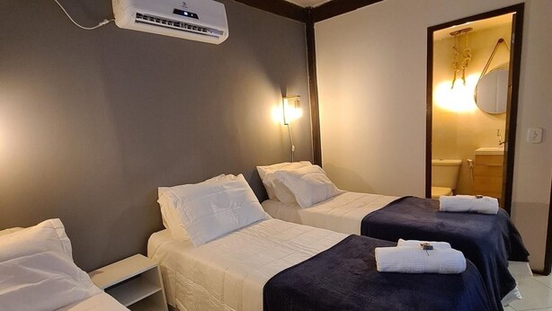 Habitación de hotel con tres camas, cada una con edredón y almohadas, una mesita de noche y un equipo de aire acondicionado en la pared.