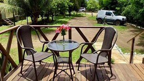 Una terraza de madera con dos sillones de mimbre y una mesa de vidrio, rodeada de exuberante vegetación y palmeras.