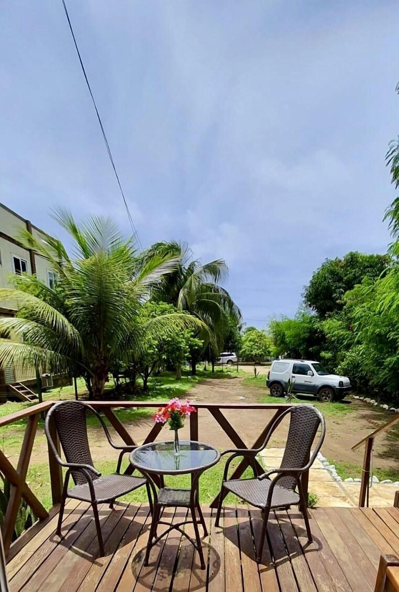 A wooden deck with two wicker chairs and a glass table, surrounded by lush greenery and palm trees.