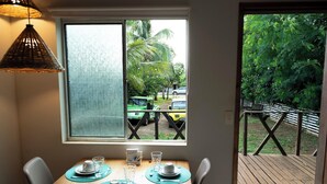 Un comedor con una mesa preparada para dos, una ventana que da a un vehículo y follaje, y una terraza de madera en el exterior.