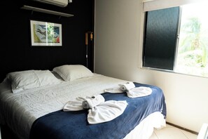 A neatly made bed with a blue blanket and white pillows, a framed artwork on the wall, and a window with a view of greenery. - Estrela Cordel (Fernando de Noronha)