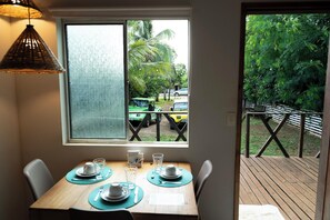 Un coin repas avec une table dressée pour deux personnes, une fenêtre donnant sur un véhicule et sur la verdure, et une terrasse en bois à l’extérieur.