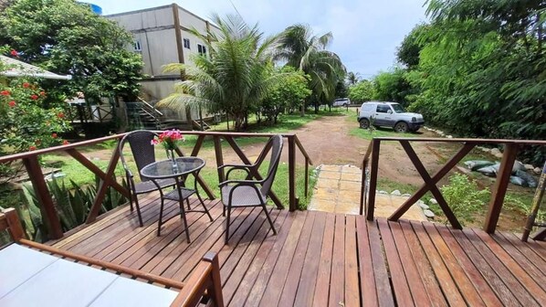 Une terrasse en bois avec deux tables et des chaises, entourée d’une végétation luxuriante et un chemin menant à un véhicule garé.
