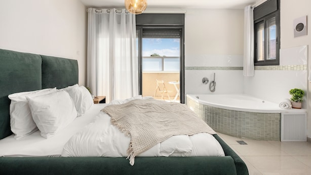 Un dormitorio moderno con cabecera verde, ropa de cama blanca, una lámpara colgante y vistas a un balcón con una silla.