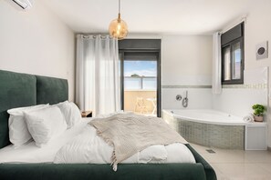 Quarto moderno com cabeceira verde, roupa de cama branca, luminária pendente e vista para uma varanda com uma cadeira.