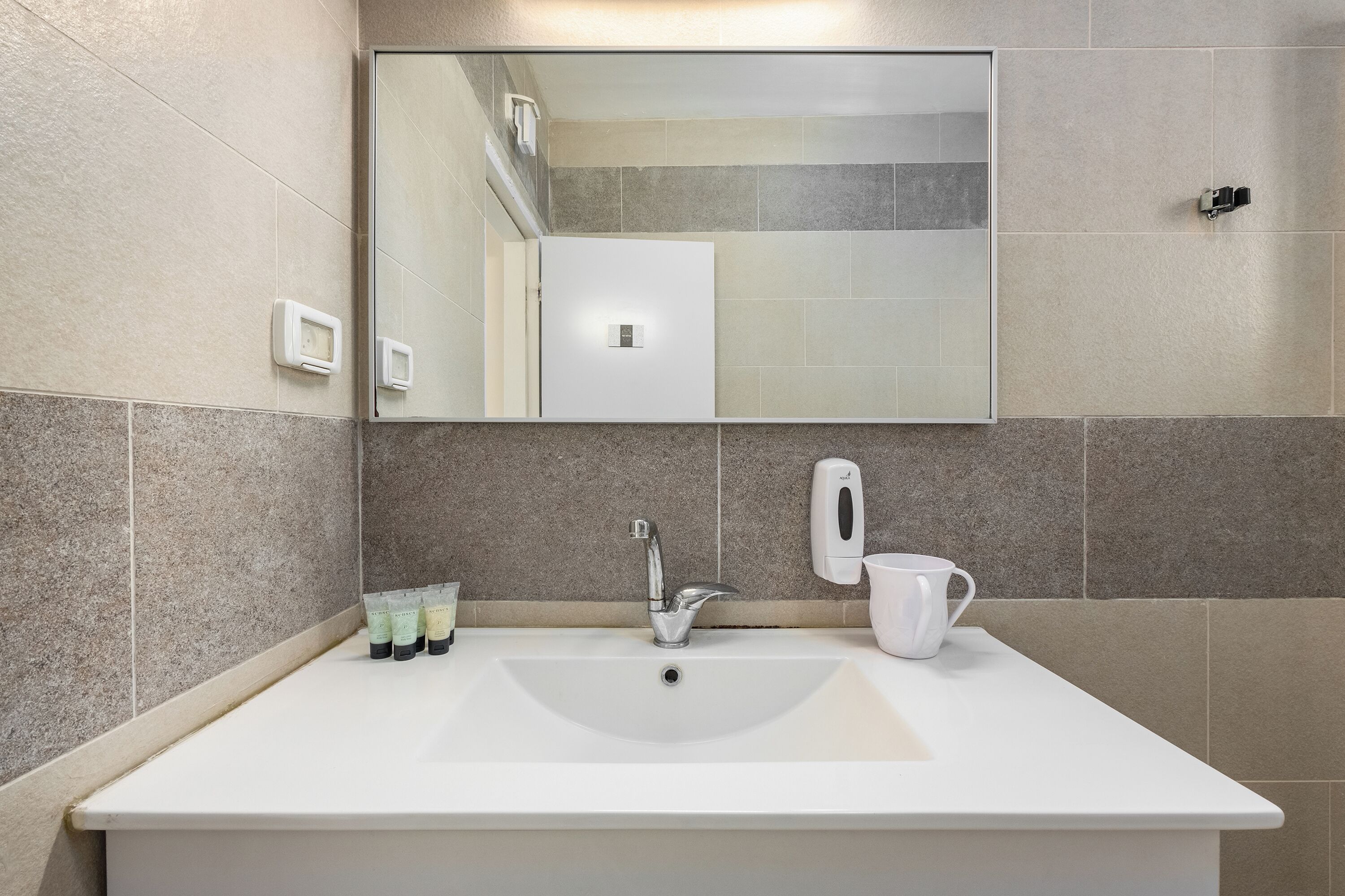 Un baño con un espejo grande, un lavamanos blanco, un dispensador de jabón y una taza.