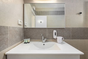 Un baño con un espejo grande, un lavamanos blanco, un dispensador de jabón y una taza.