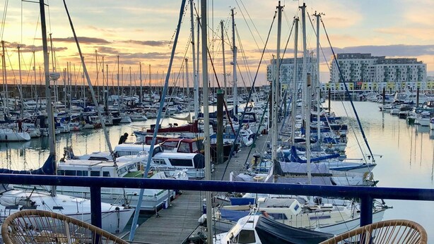 Un puerto deportivo con numerosos barcos amarrados, cielo despejado y vista de un horizonte urbano al fondo.