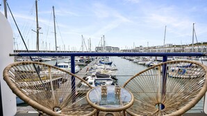 Un balcón con sillones de mimbre y una mesa de vidrio con vistas a un puerto deportivo con barcos y edificios.