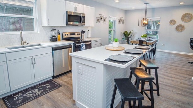 Une cuisine moderne avec des meubles blancs, un îlot central doté de tabourets et un coin repas avec une longue table.