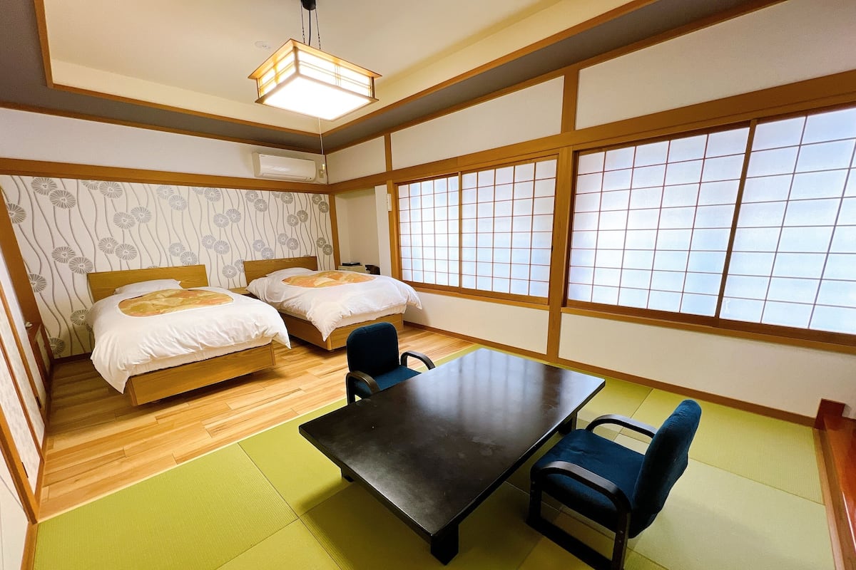 Quarto em estilo japonês tradicional, com tatames, duas camas de madeira, uma mesa e vista através de biombos shoji.