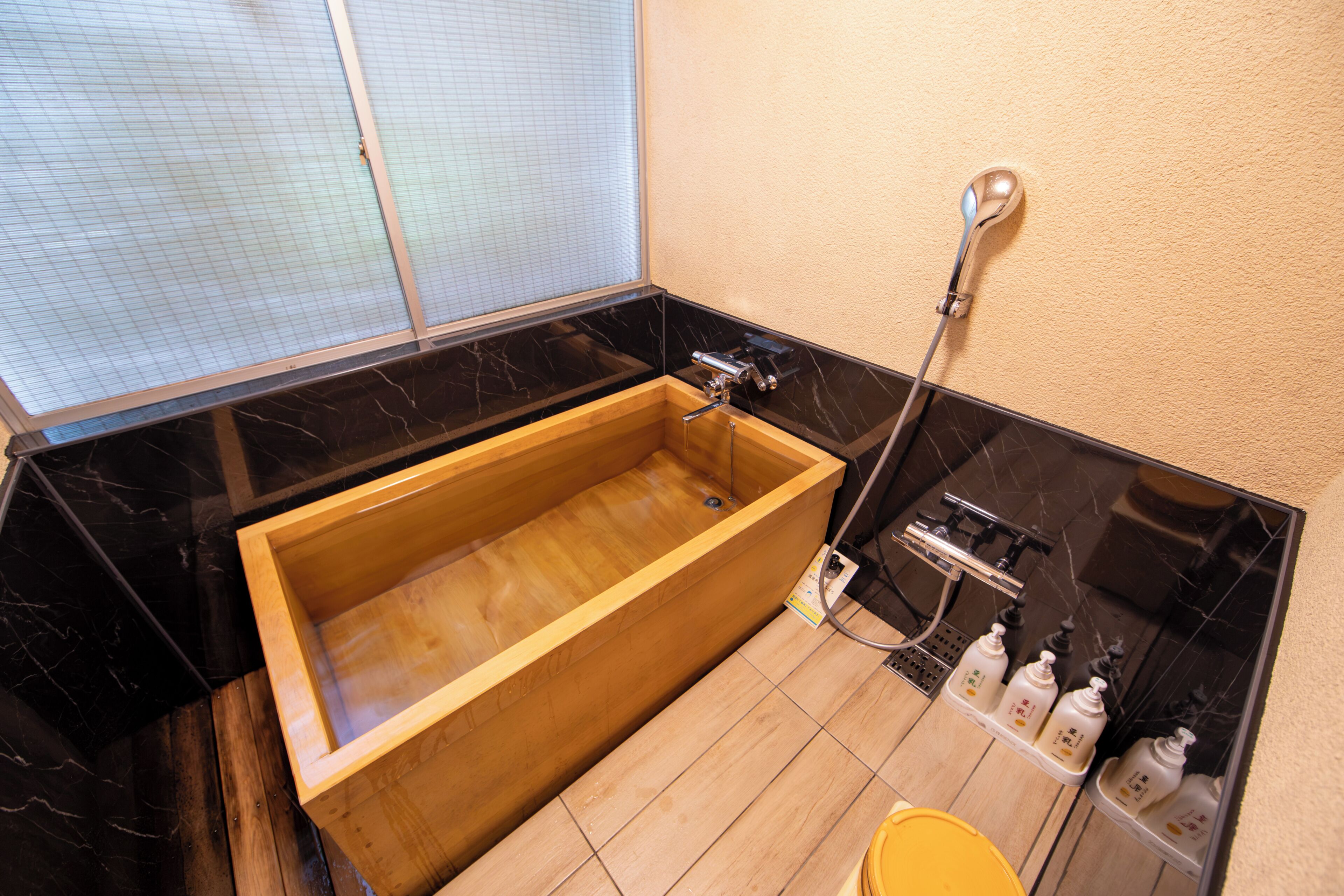A wooden bathtub in a bathroom with a black marble countertop, a shower head, and toiletries on the side.