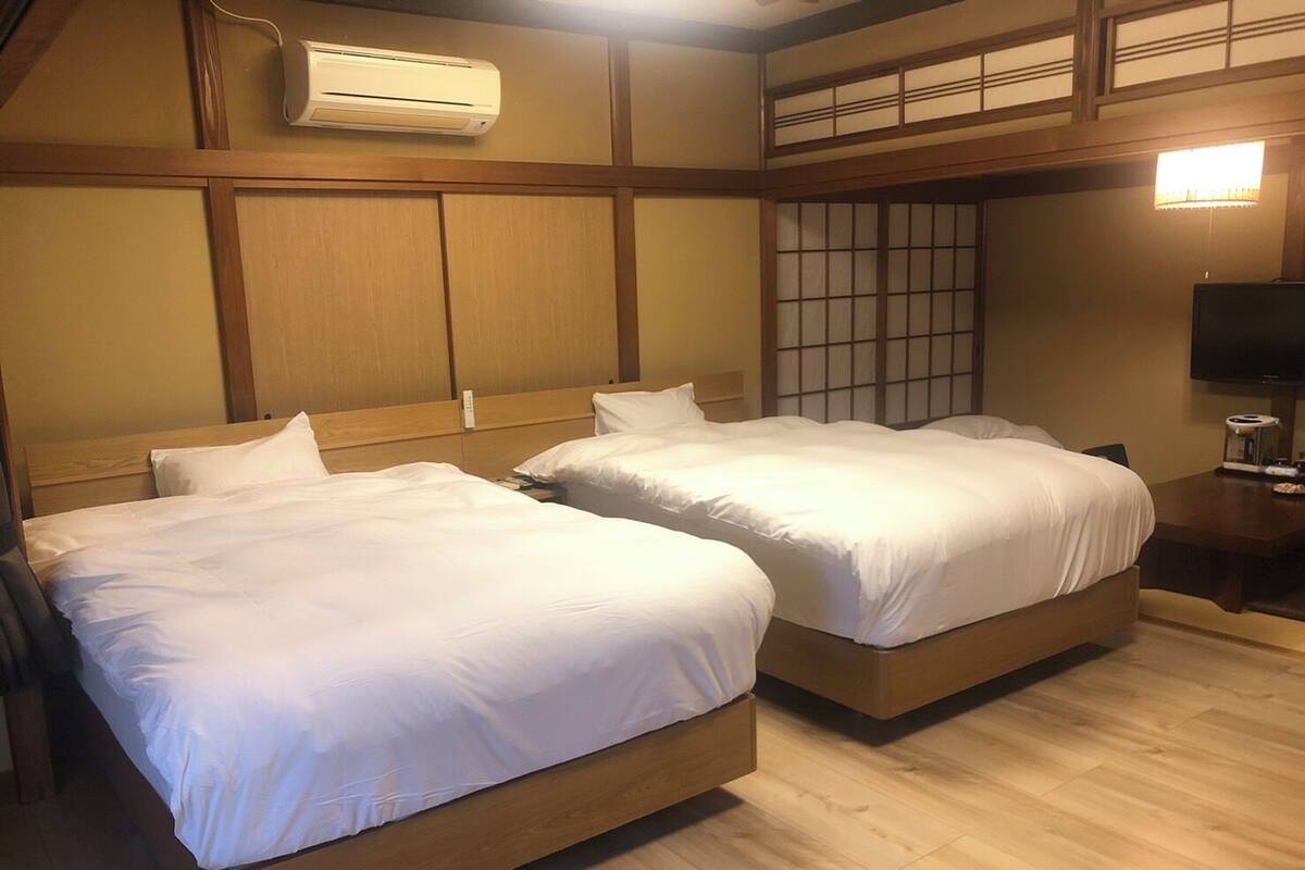 Quarto de hotel com duas camas de madeira, ar condicionado embutido, um tradicional divisória japonesa shoji e uma televisão sobre uma mesa de madeira.