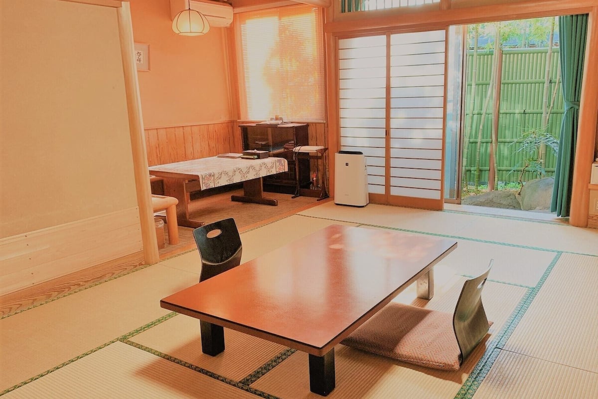 Quarto em estilo japonês tradicional, com tatames, uma mesa baixa, uma cadeira e uma porta deslizante para um jardim de bambu.