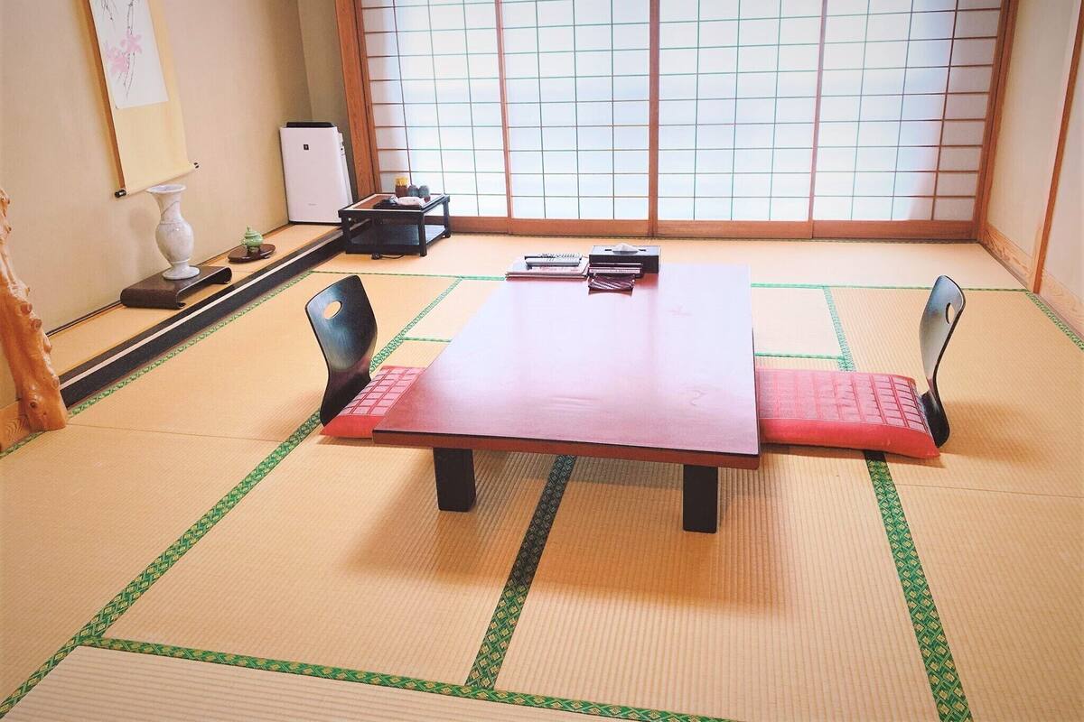 Um espaço com esteiras de tatame, uma mesa de madeira, duas cadeiras, uma pequena escrivaninha com um computador e uma janela com biombos shoji.