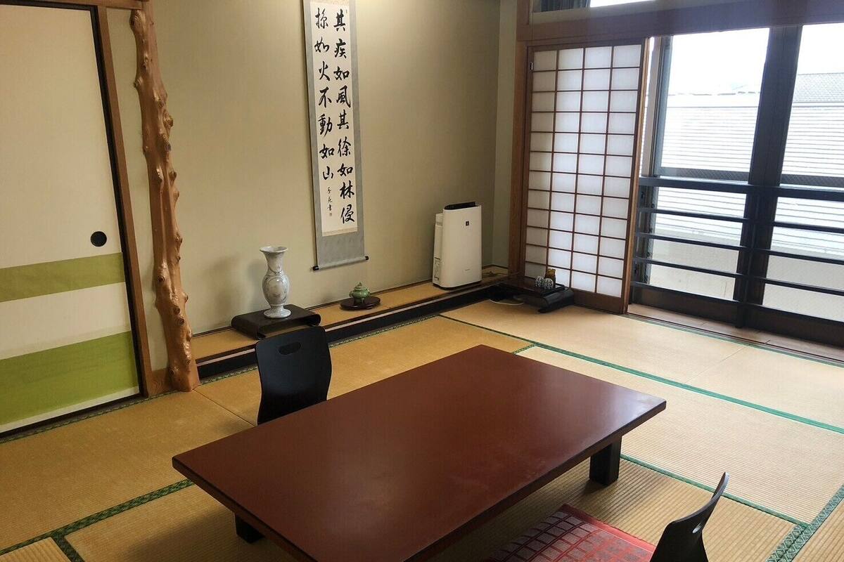 Um quarto japonês tradicional com tatames, uma mesa baixa, uma cadeira, um painel shoji e um pergaminho suspenso com caligrafia japonesa.