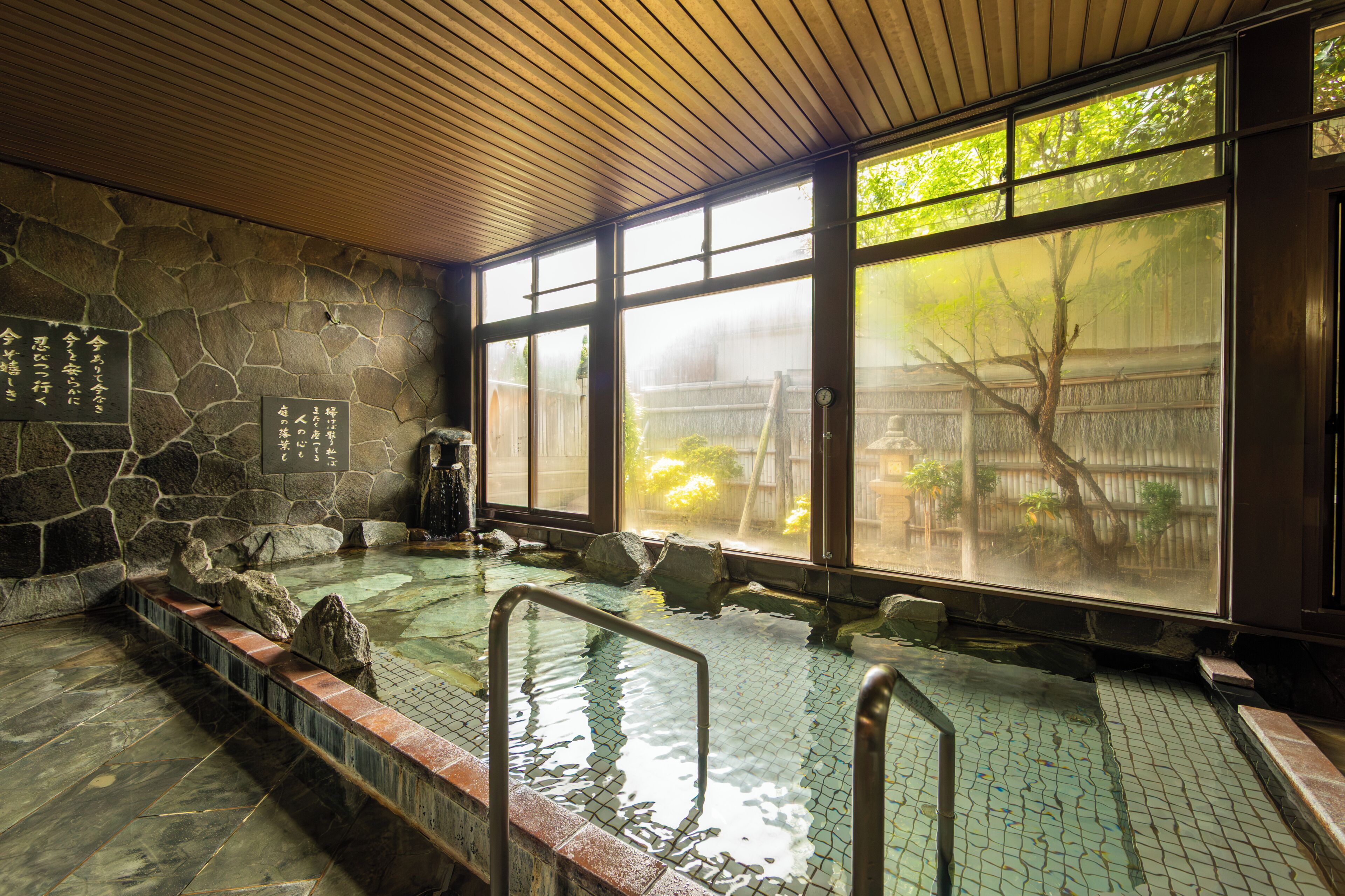 A traditional Japanese onsen with stone walls, a tiled floor, and a view of a garden through large windows.