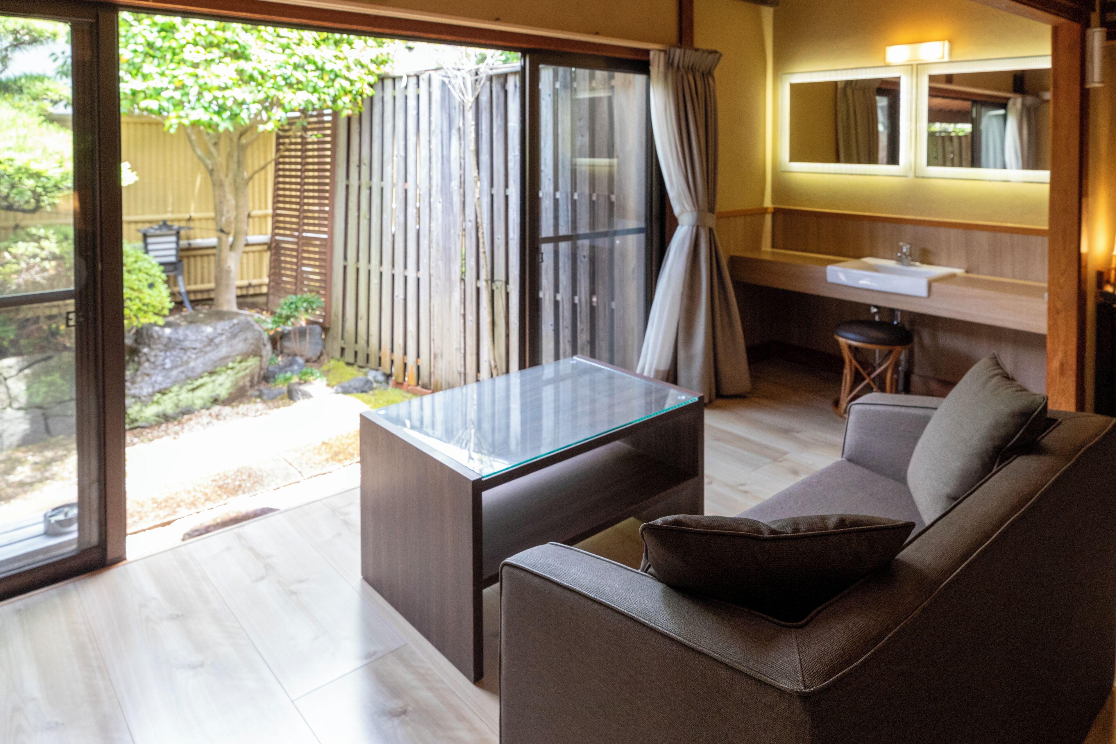 A modern interior with a glass-top table, a sofa, a bathroom with a sink and mirror, and a view of the outdoors.